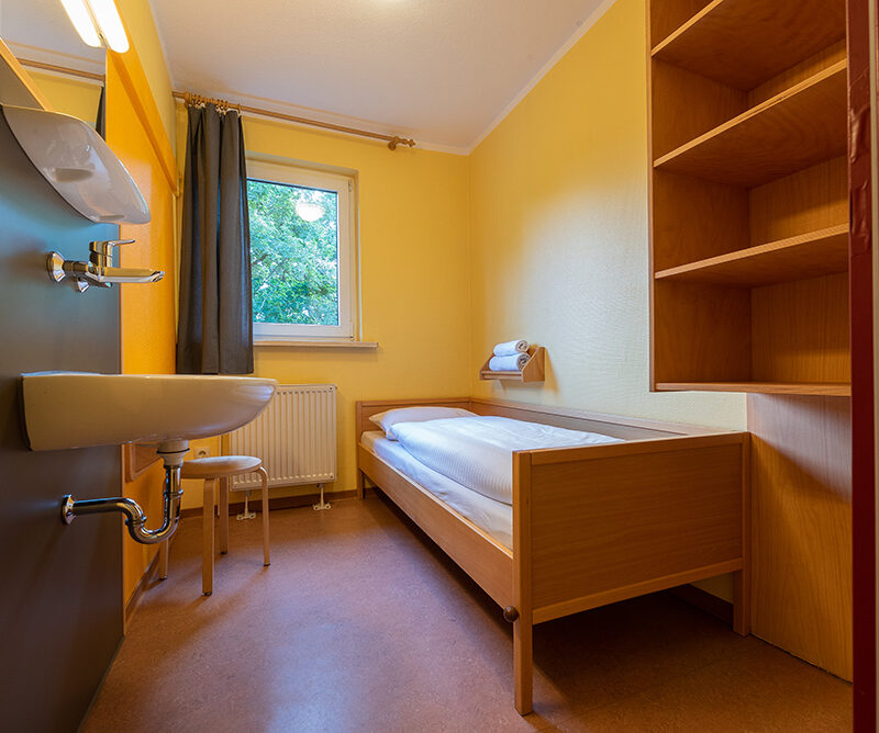 Einzelzimmer mit gemachtem Bett, einer Ablage mit Handtüchern, einem Hocker, Fenster. Im Vordergrund links ein Waschbecken, rechts ein Regal.