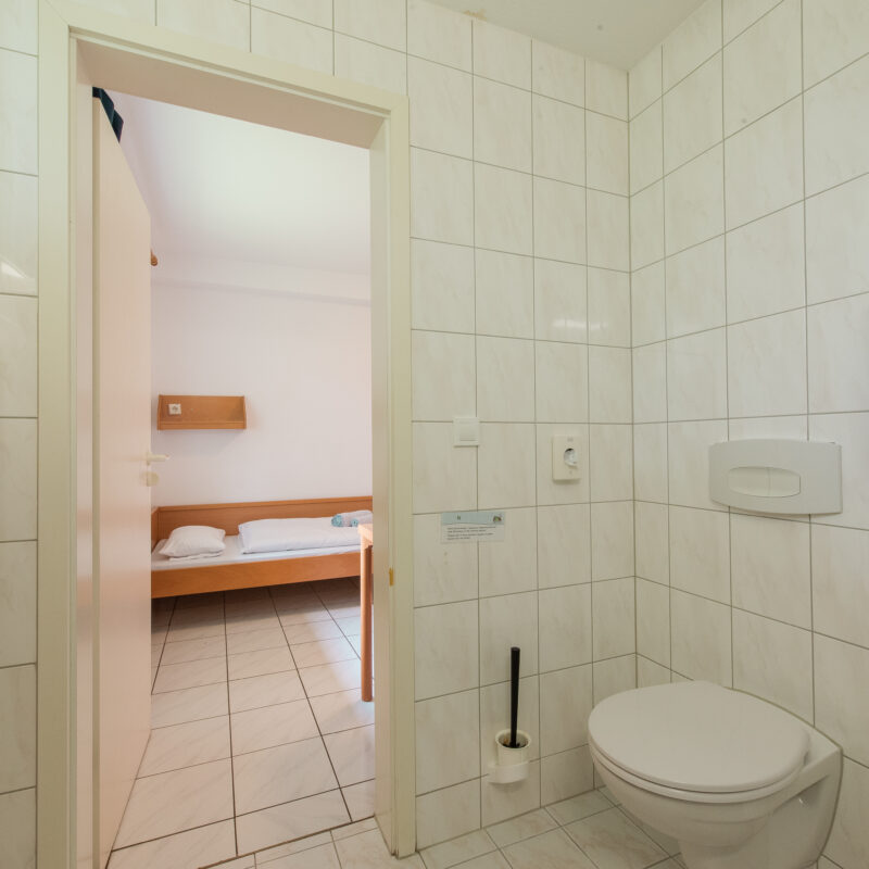 Blick vom privaten Bad mit Toilette ins Zimmer. Im Zimmer befindet sich ein Bett. Das ist bezogen und Handtücher liegen als Rolle drauf.