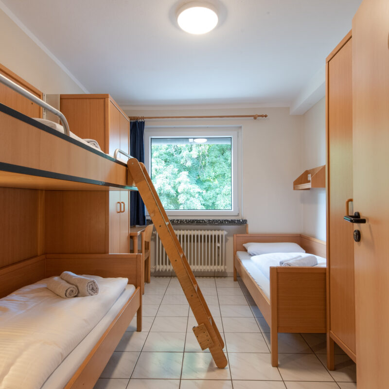 Blicl in ein 3-Bettzimmer mit links einem Etagenbett. Leiter steht am Hochbett an. Rechts hinter dem Schrank ein einzelstehendes Bett. Blick weiter aus dem Fenster auf einen grünen Baum.