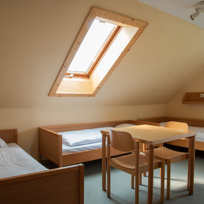 Mehrbettzimmer im Dachgeschoss mit Dachschräge und Fenster in der Dachschräge. Zu sehen sind drei gemachte Betten und ein kleiner Tisch mit zwei Stühlen in der MItte des Zimmers.