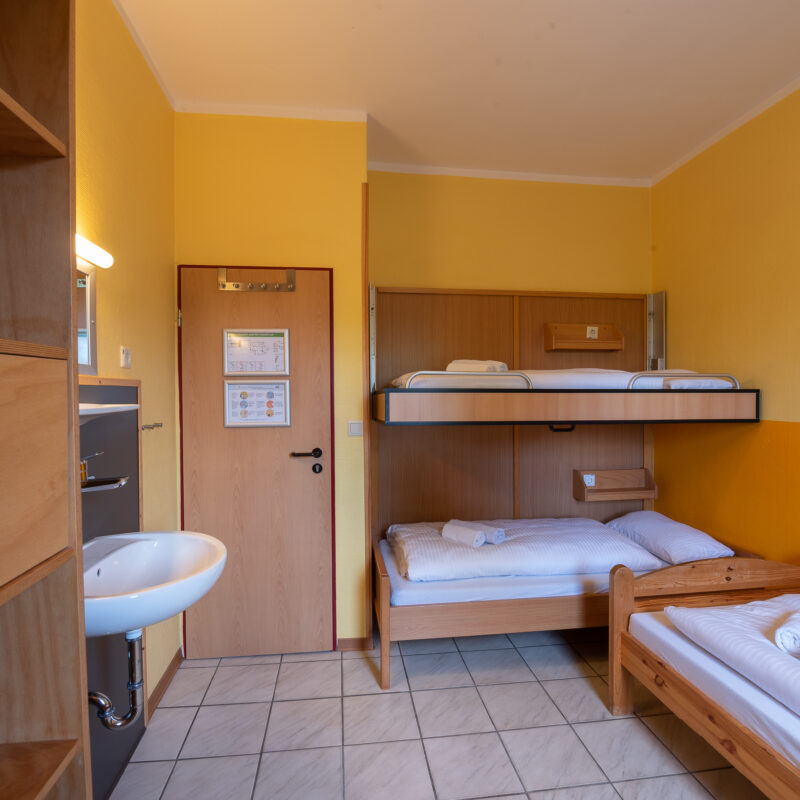 Im Zimmer sind ein Etagenbett und ein einzelstehendes Bett. Die Betten sind gemacht. Links an der Wand ein Waschbecken mit Spiegel und daneben ein Schrank aus Holz mit teilweise offenen Fächern und Fächern mit Türen.