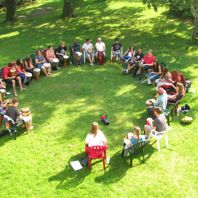 Eine Gruppe junger Erwachsener sitzen im Garten im Kreis. Sicht von oben auf die Gruppe. Alle haben eine Trommel zwischen den Beinen.