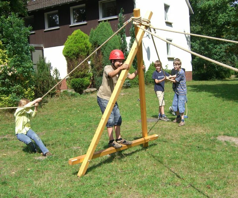 Erlebnispädagogisches Element aus Holz und in Form eines mannshohen Dreiecks wird mit vier Seilen an der Spitze von Kindern gehalten. Im Dreieck steht ein Kind mit Helm.
